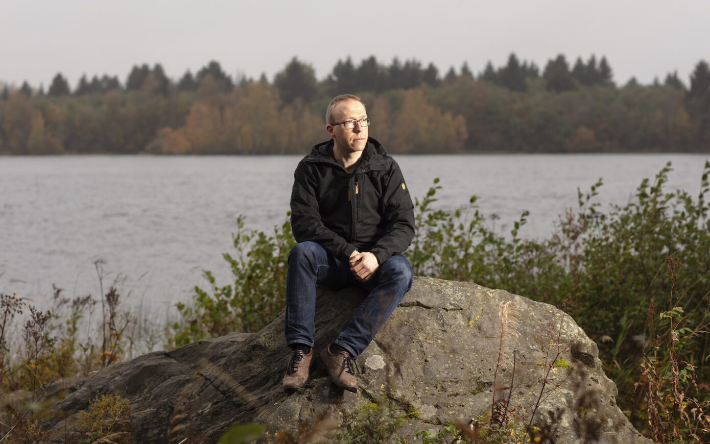 Jouni Rissanen sits on a rock and looks to his right. Behind his back is a grey sea or lake.