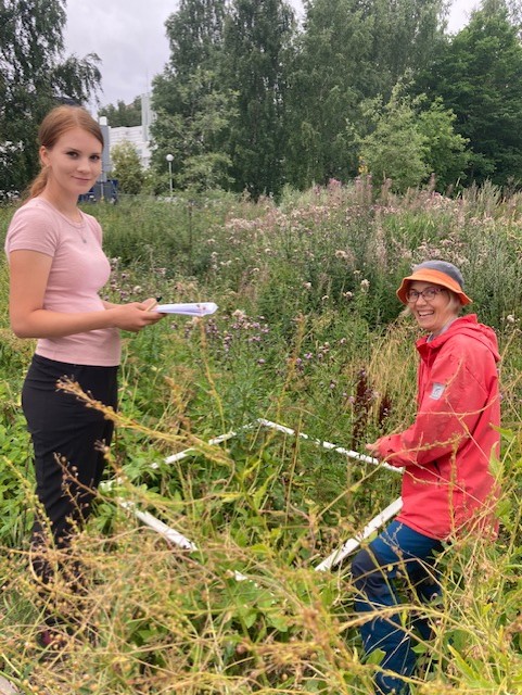 Two women in the middle of a flower field collecting samples, looking into the camera and smiling