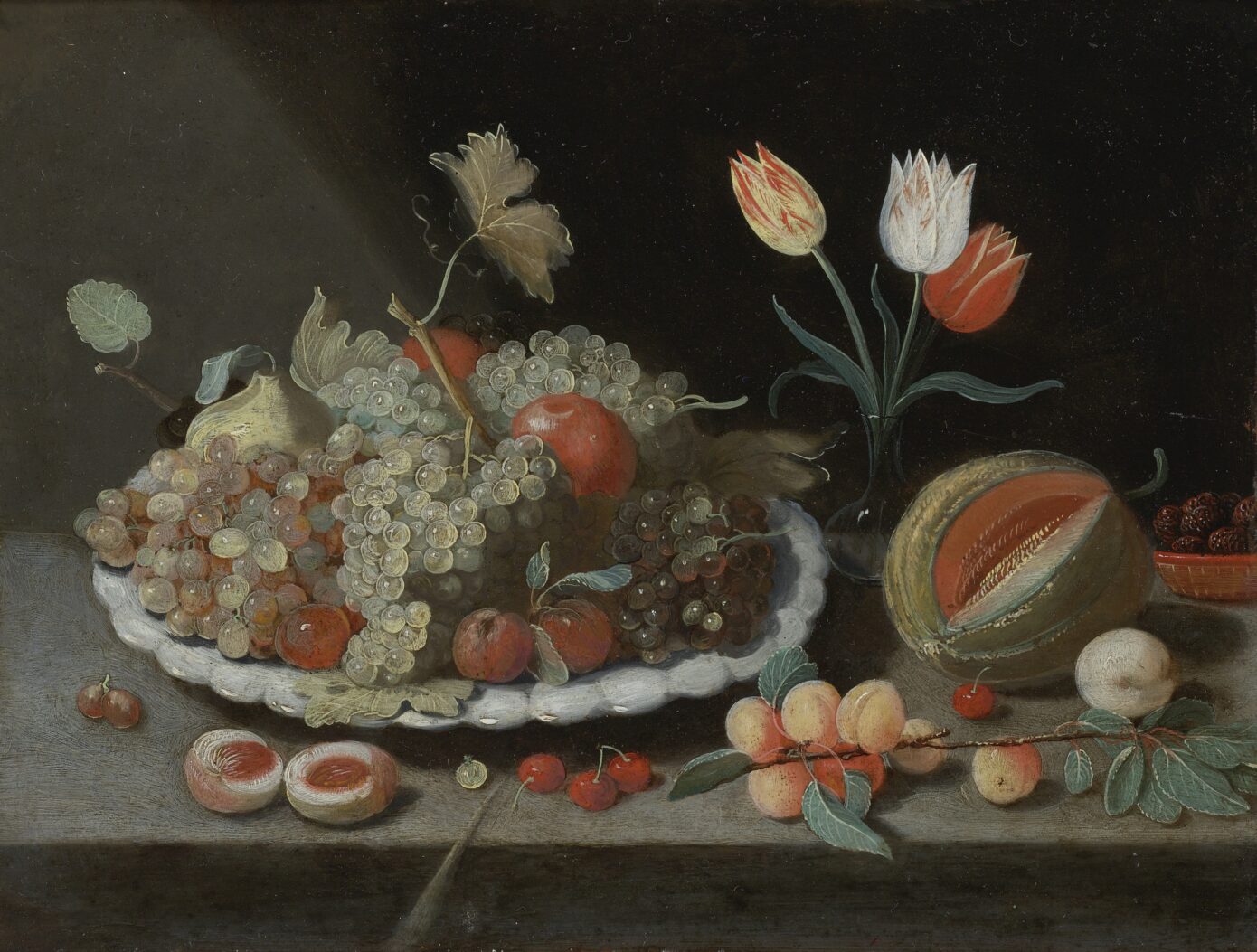 Still life with grapes and other fruit on a platter, a glass vase with tulips, a melon, apricots, cherries and other fruits, all on a ledge